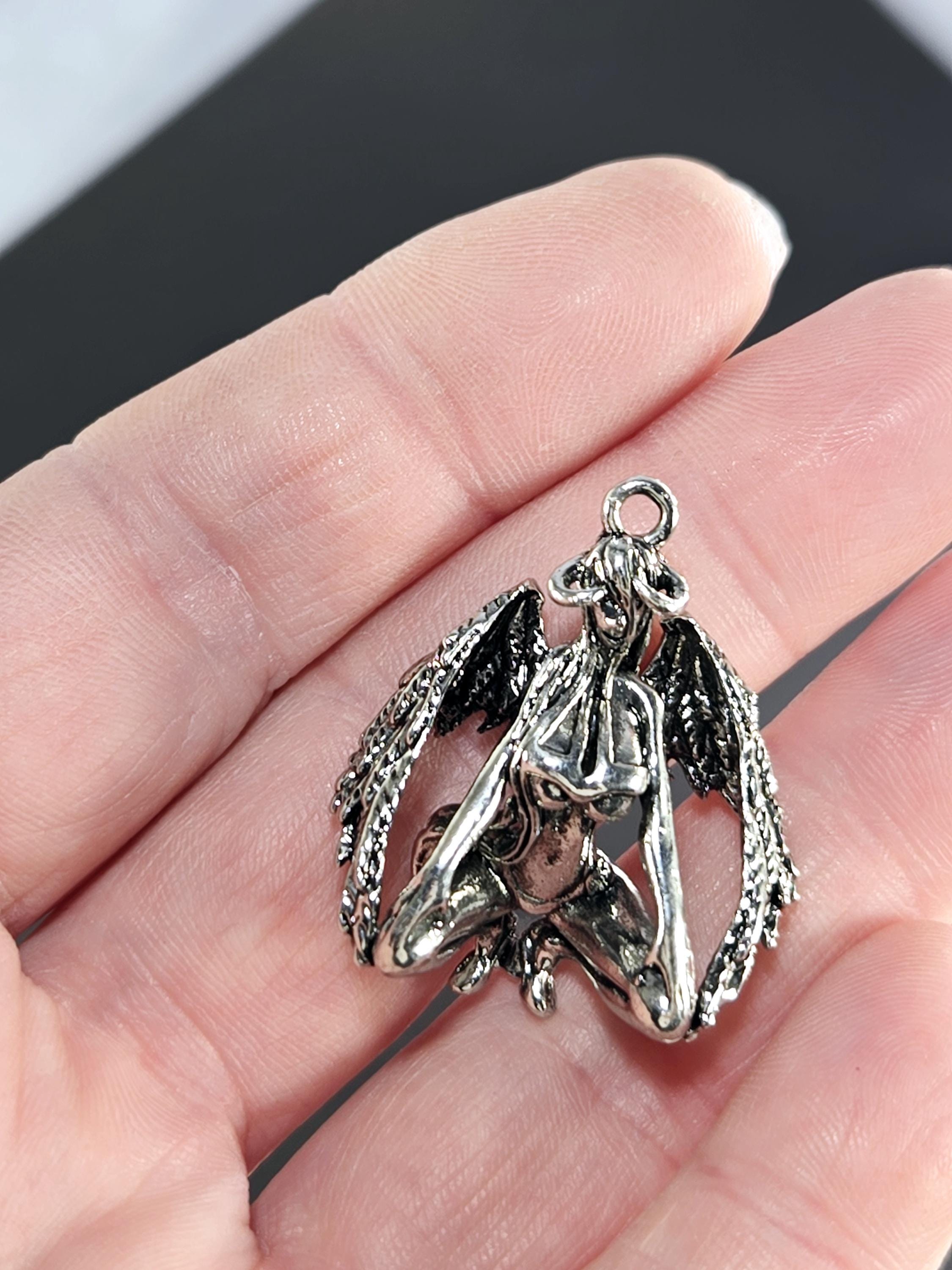A close-up image of a person's hand holding a silver-colored pendant or charm in the shape of a bird or winged figure.