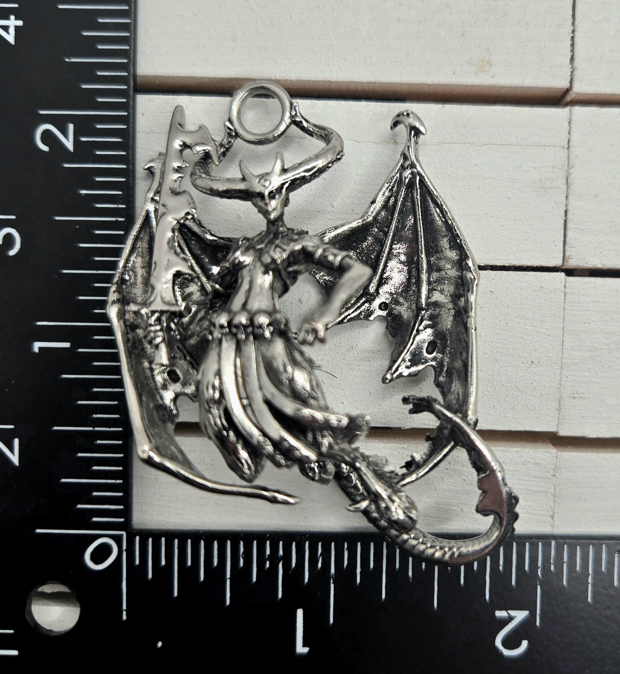 A silver-colored metal pendant in the shape of a dragon with wings and a tail, resting on a ruler for size comparison.