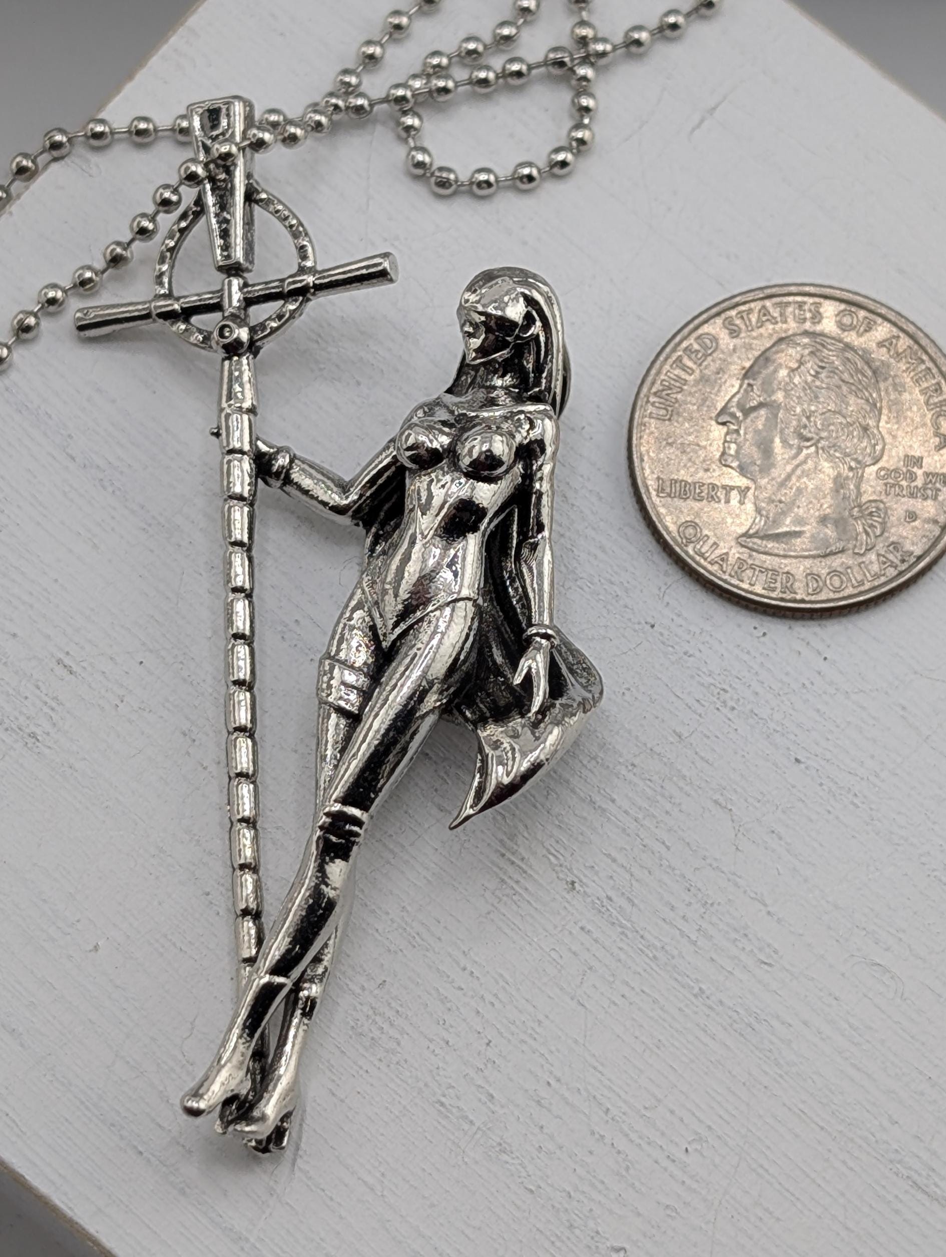 A silver-colored pendant in the shape of a woman holding a cross, placed next to a United States quarter coin.