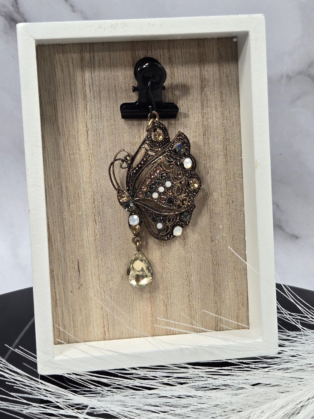 A decorative butterfly pendant with a large gemstone at the center, displayed in a white ornate frame.