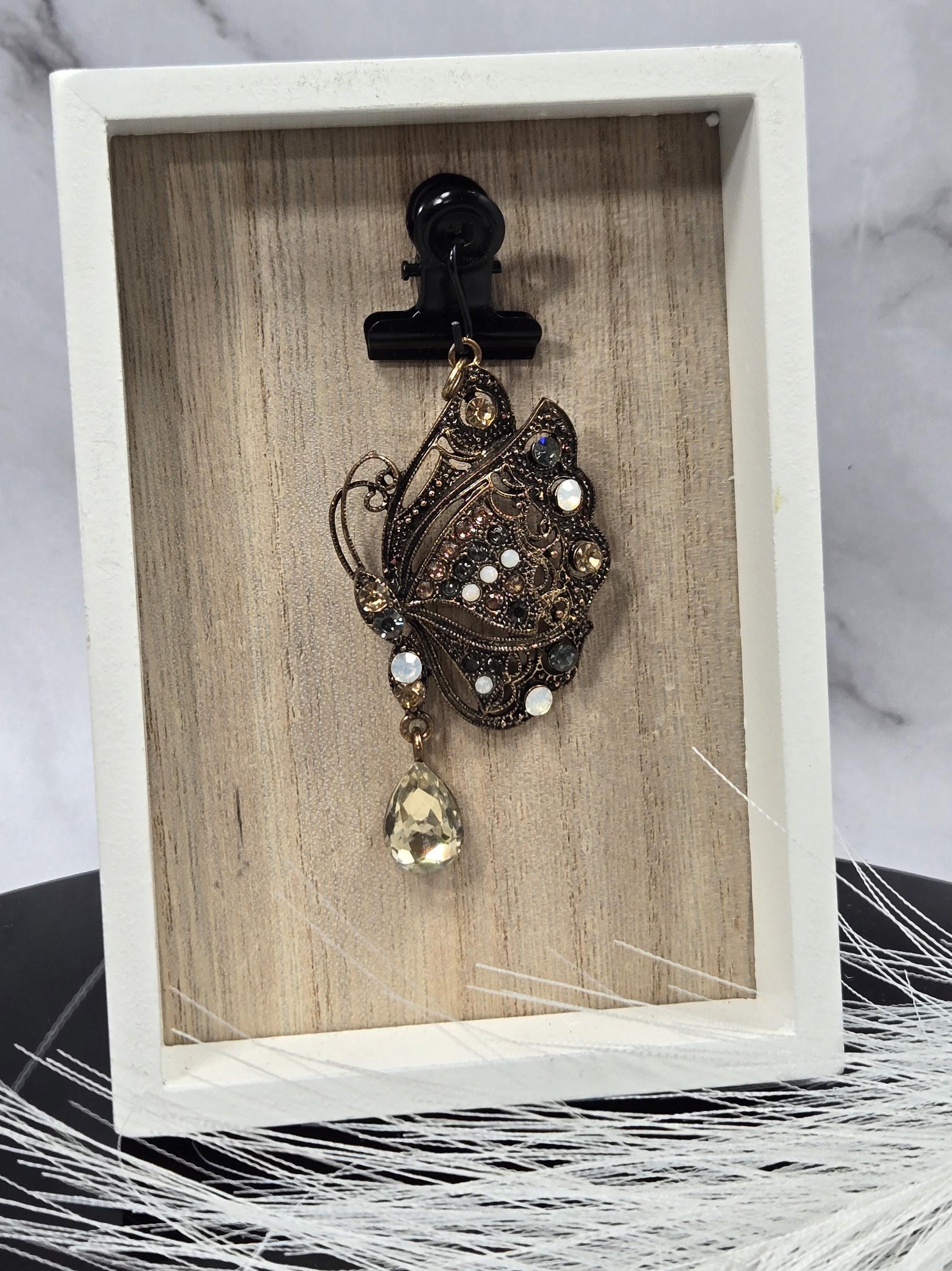 A decorative pendant with a floral design and a large gemstone is displayed in a white wooden frame.