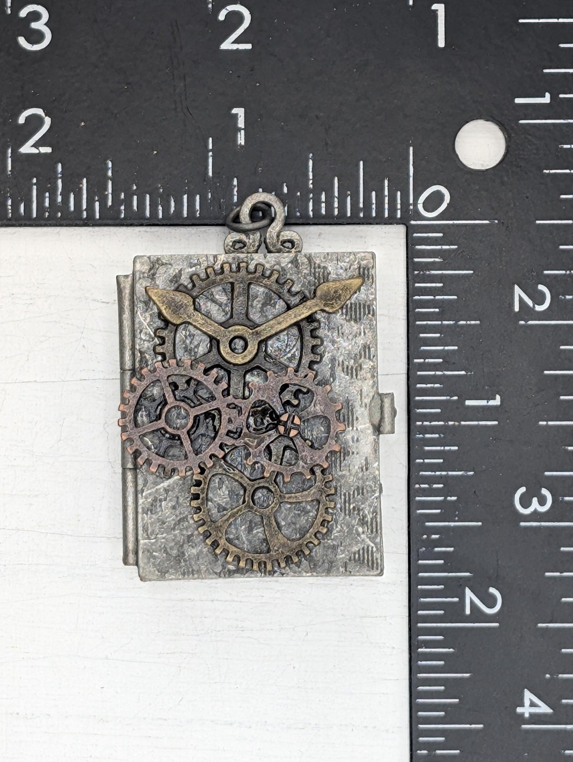A metal pendant in the shape of a book with gears and cogs attached to it, placed on a ruler for size comparison.