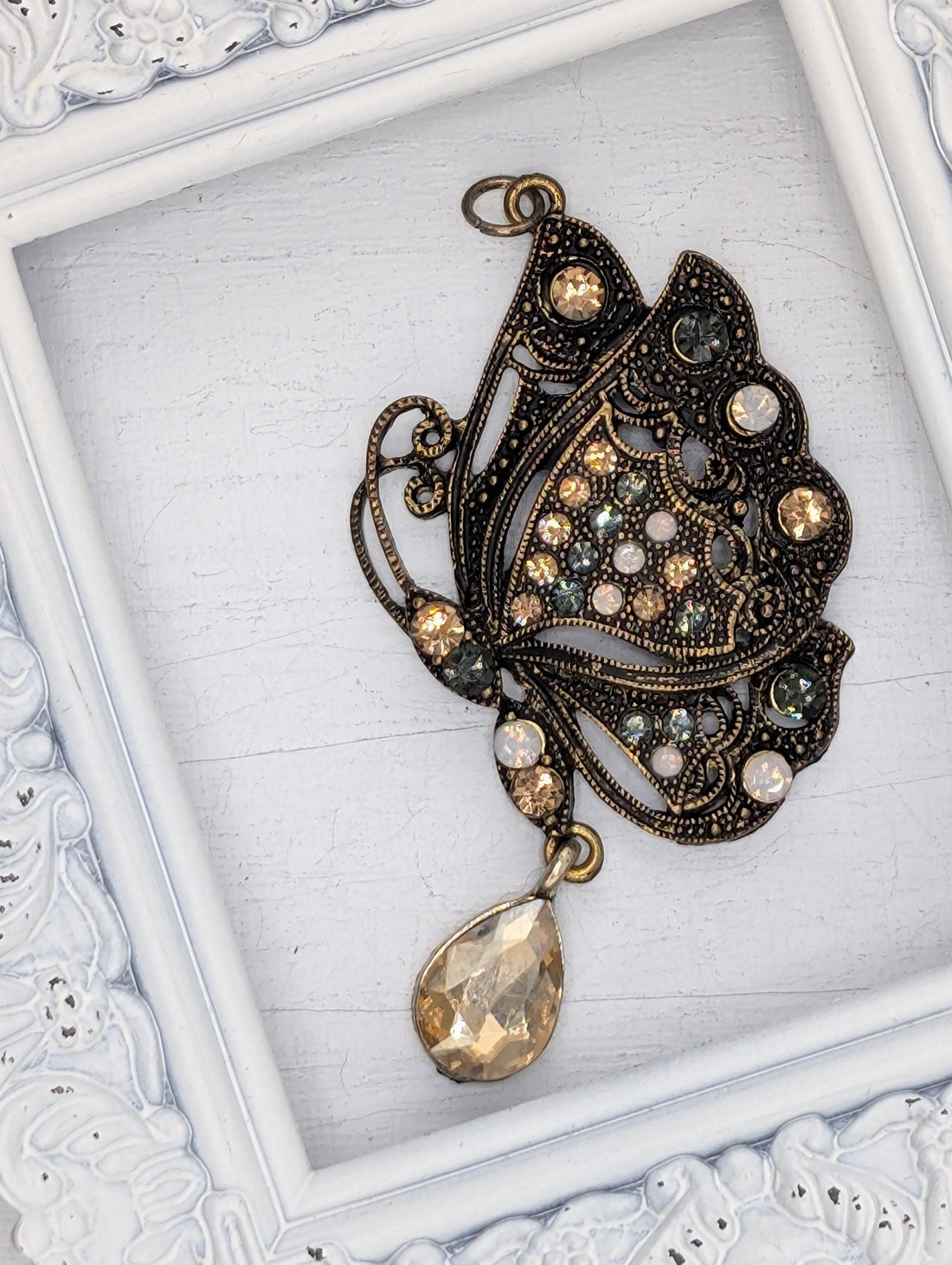 A decorative butterfly pendant with a large gemstone at the center, displayed in a white ornate frame.