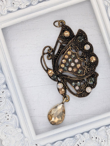 A decorative butterfly pendant with a large gemstone at the center, displayed in a white ornate frame.