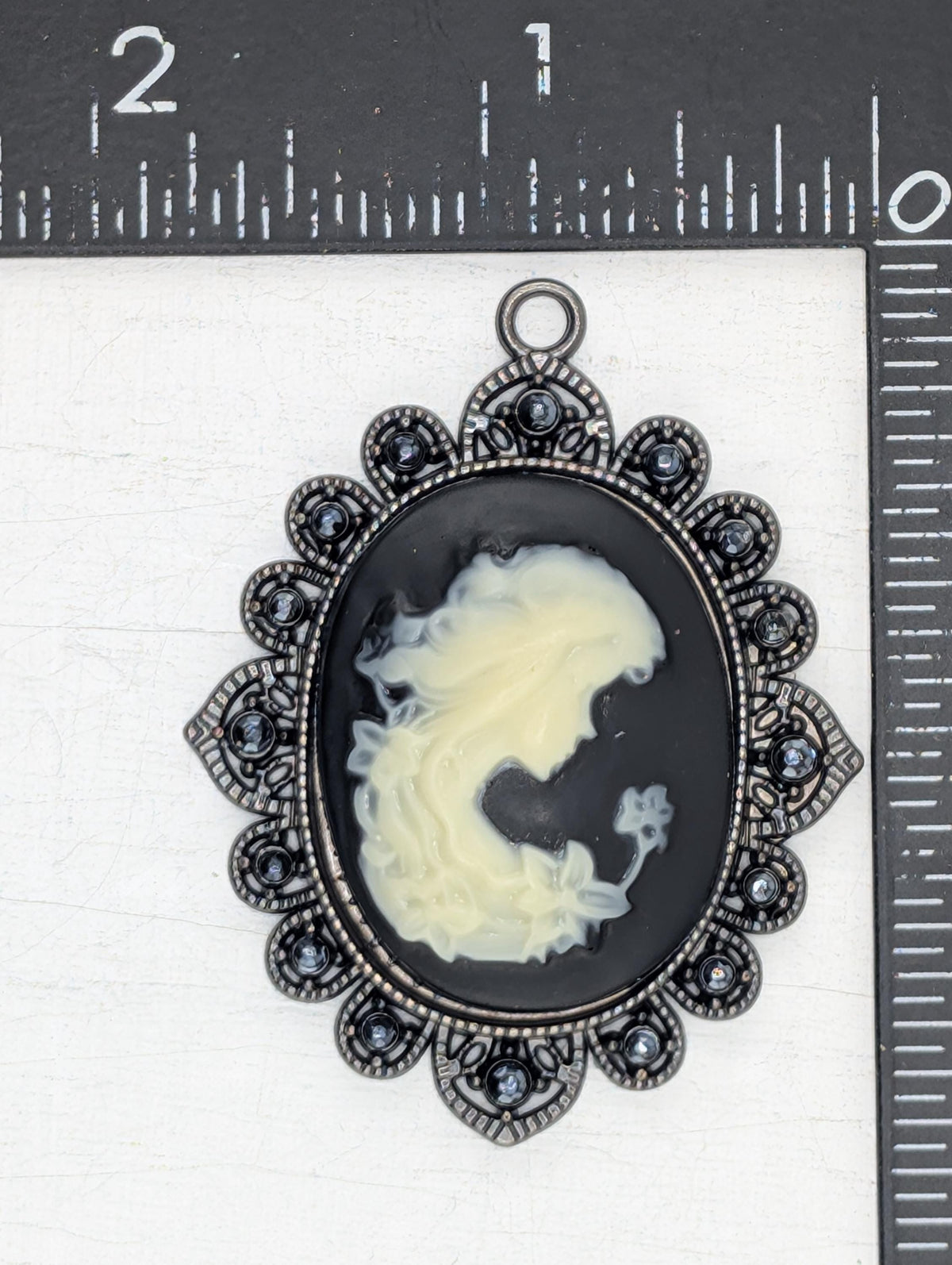 a pendant with a black oval background and a white silhouette of a woman's face. The pendant is adorned with a decorative metal frame and has small blue stones or crystals embedded in it. The pendant is placed on a white surface, and there is a ruler visible in the background.