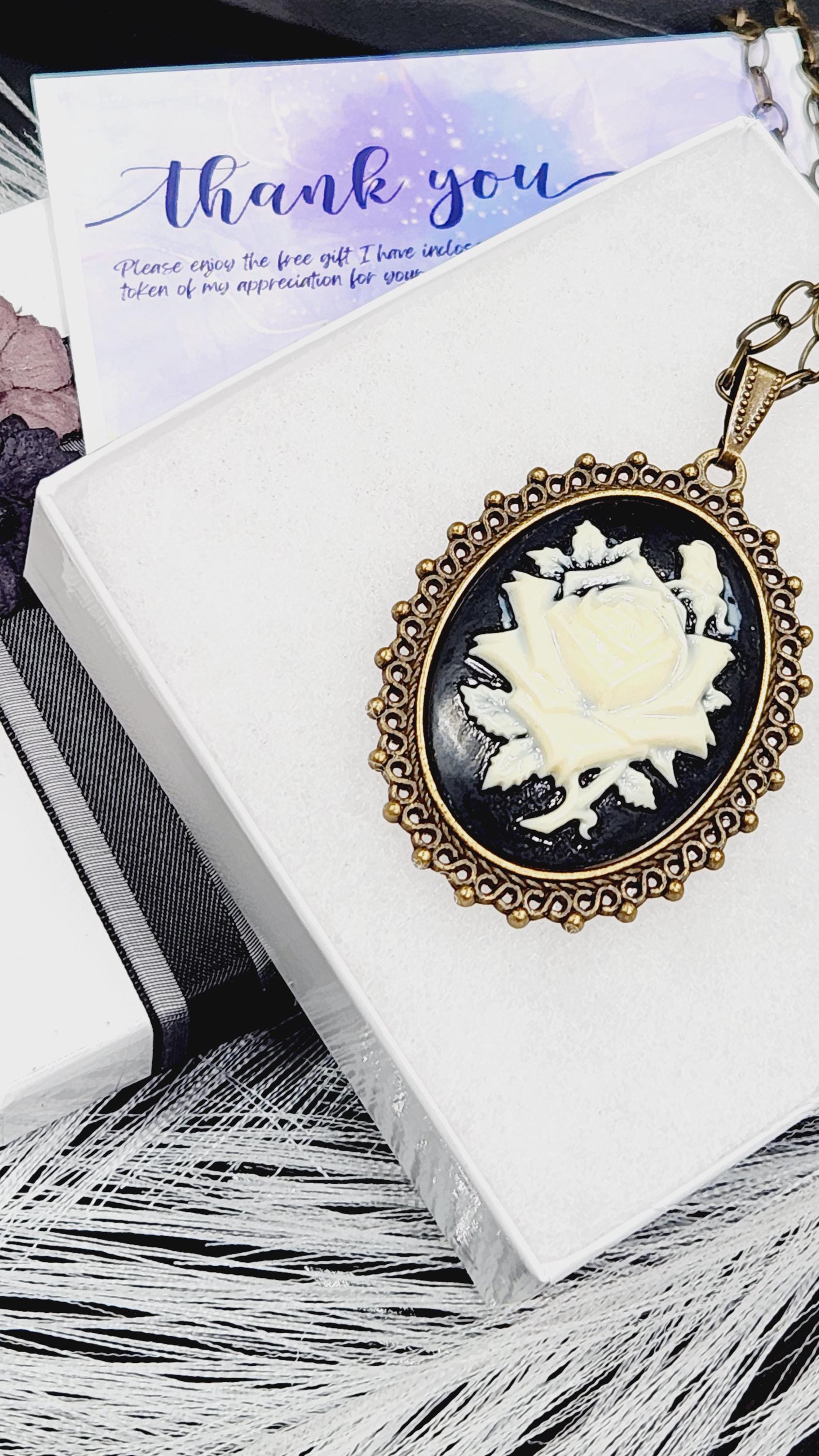 A pendant with a white rose design is displayed on a white surface, accompanied by a purple card with a "Thank you" message.