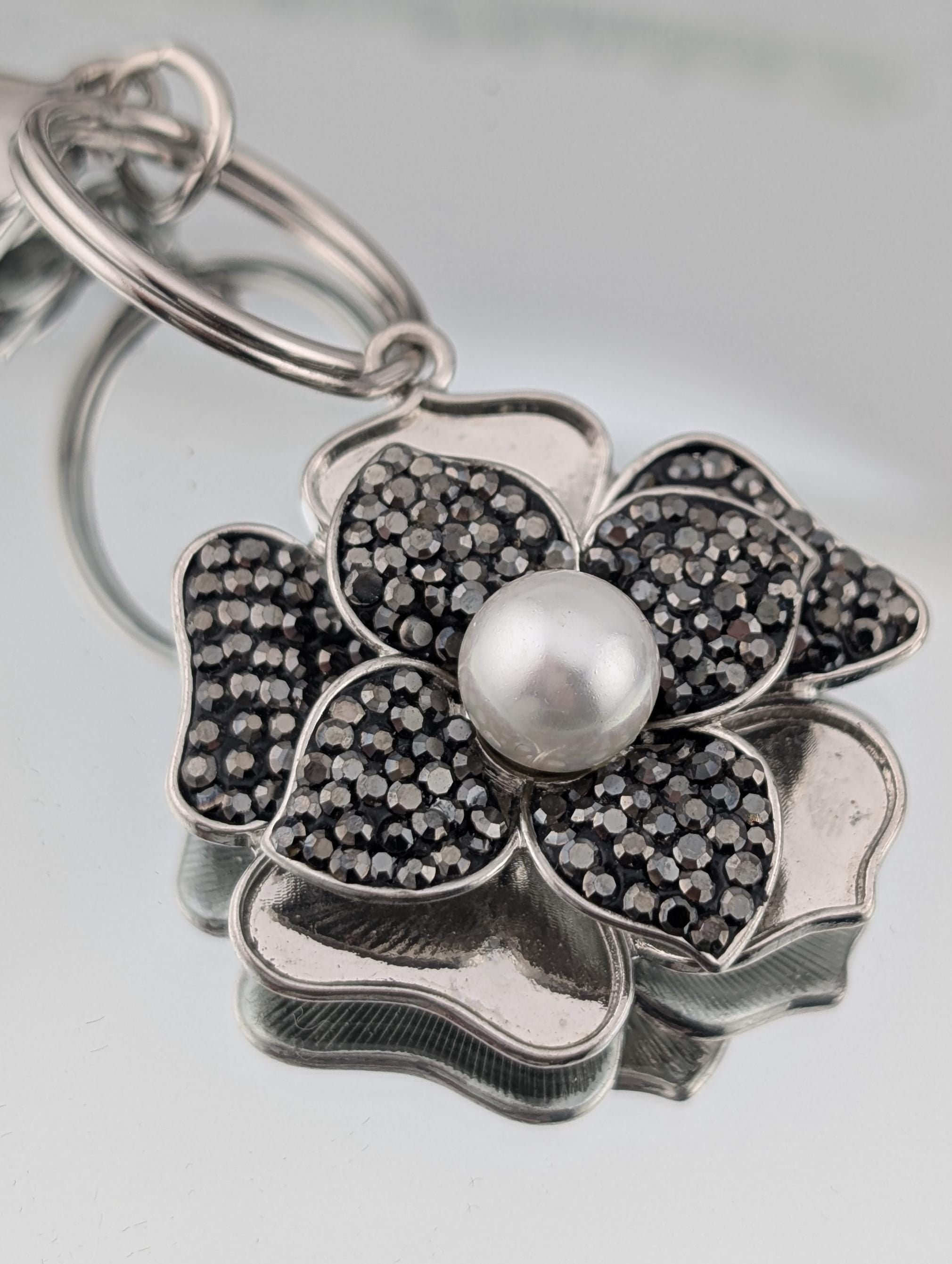 A silver-colored keychain with a flower-shaped pendant featuring a pearl at the center, surrounded by small black gemstones.
