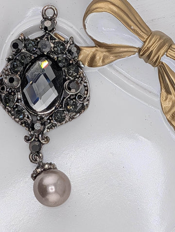 A decorative pendant with a large crystal and a pearl hanging from it, accompanied by a gold ribbon.