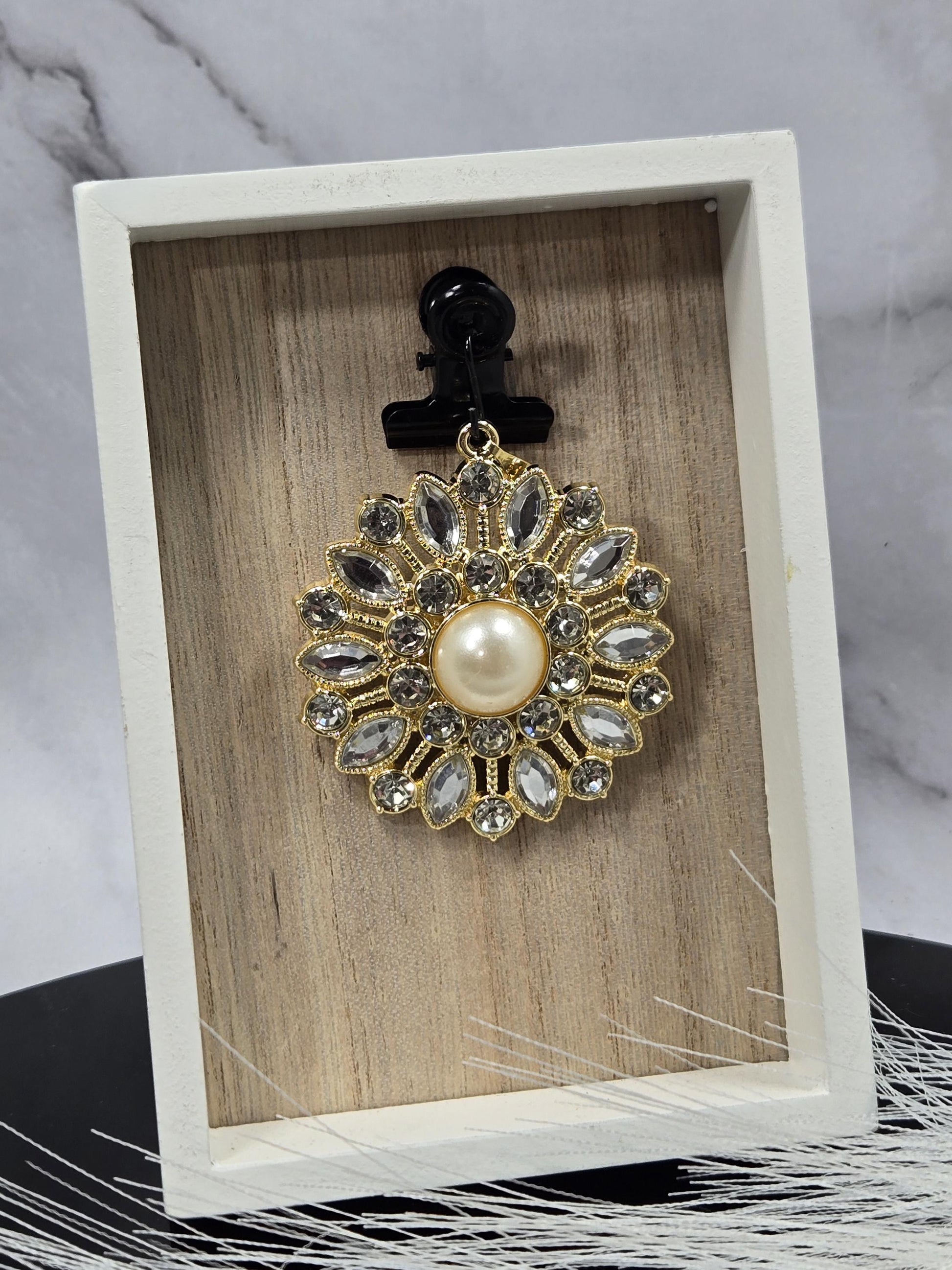 A decorative brooch with a pearl at its center, surrounded by numerous sparkling gemstones, is displayed in a white wooden box.