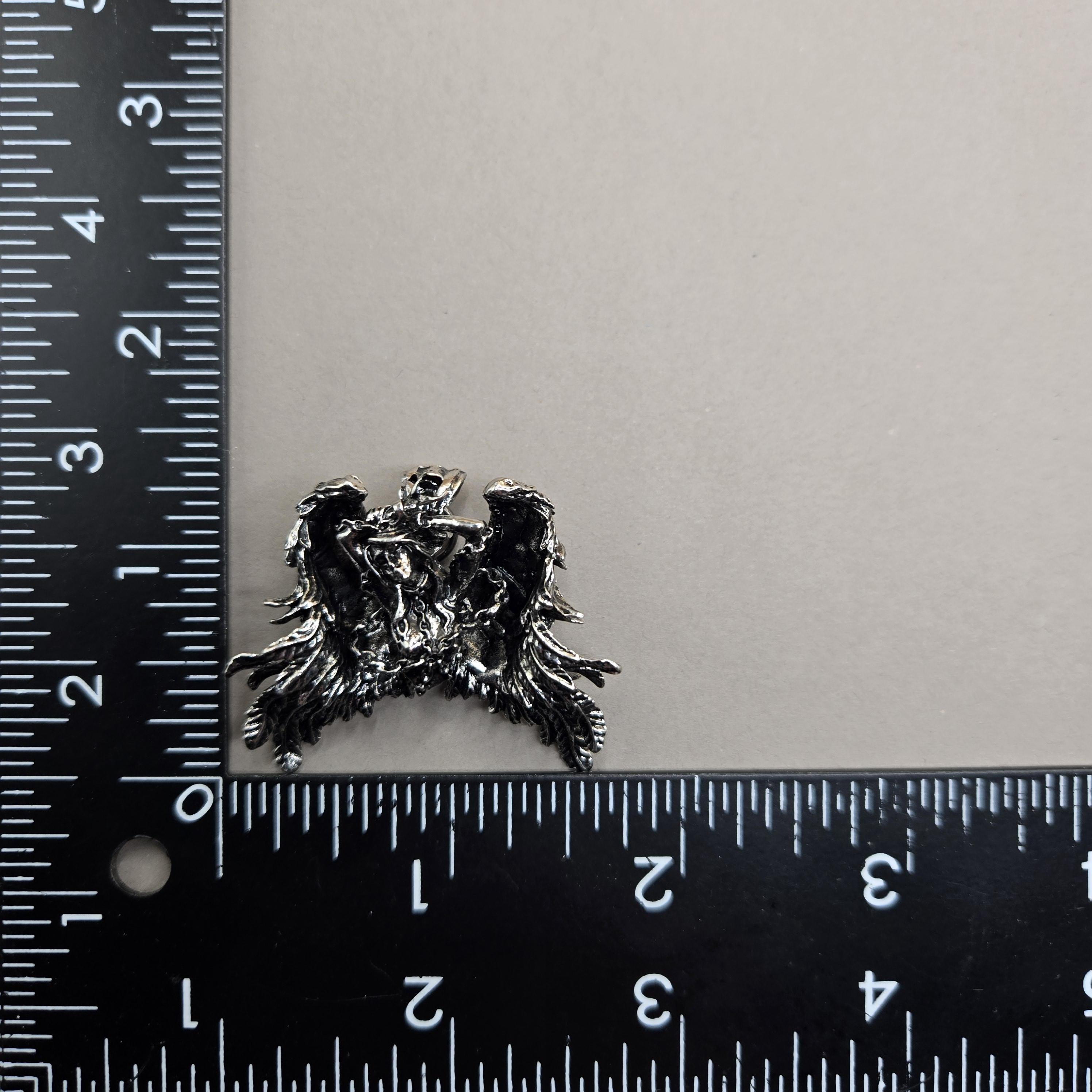 A silver-colored metal object, possibly a pendant or sculpture, is placed next to a ruler on a white surface.