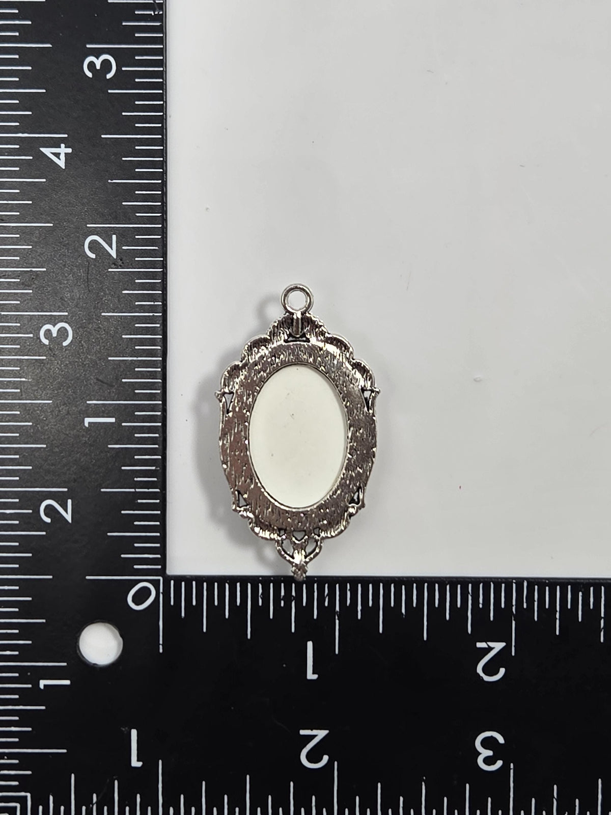 a small, ornate metal piece with a white oval-shaped center, placed next to a ruler for size comparison.
