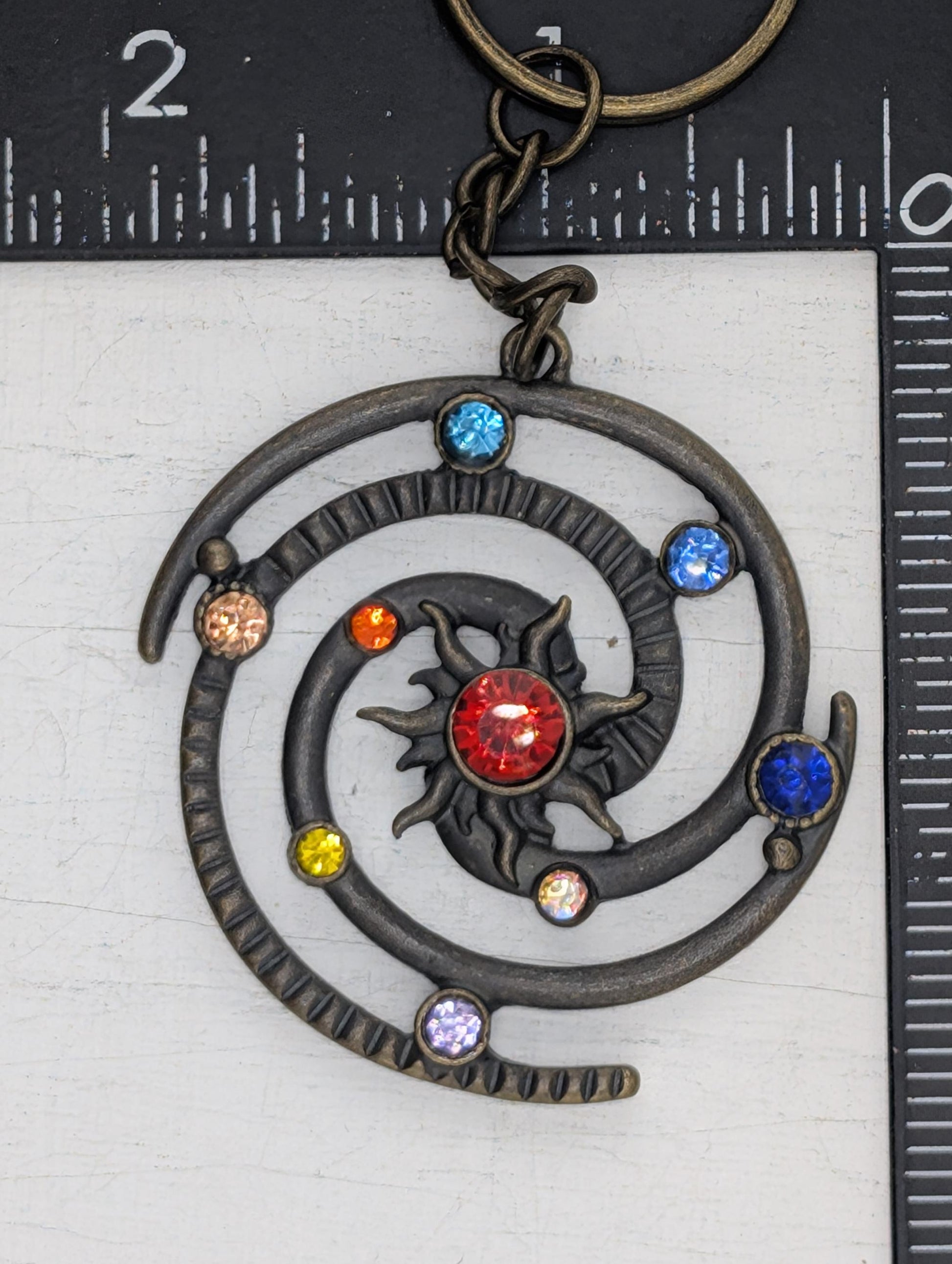 A decorative metal pendant with a spiral design and colorful gemstones, including a red gemstone at the center.