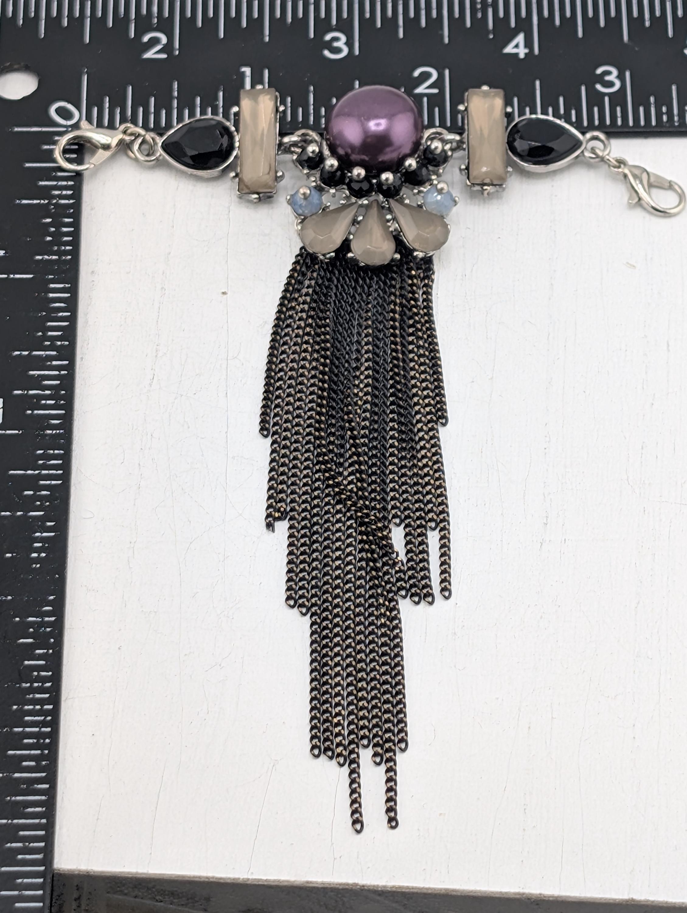 A piece of jewelry with a purple gemstone, silver chain, and black tassels, placed on a ruler for size comparison.