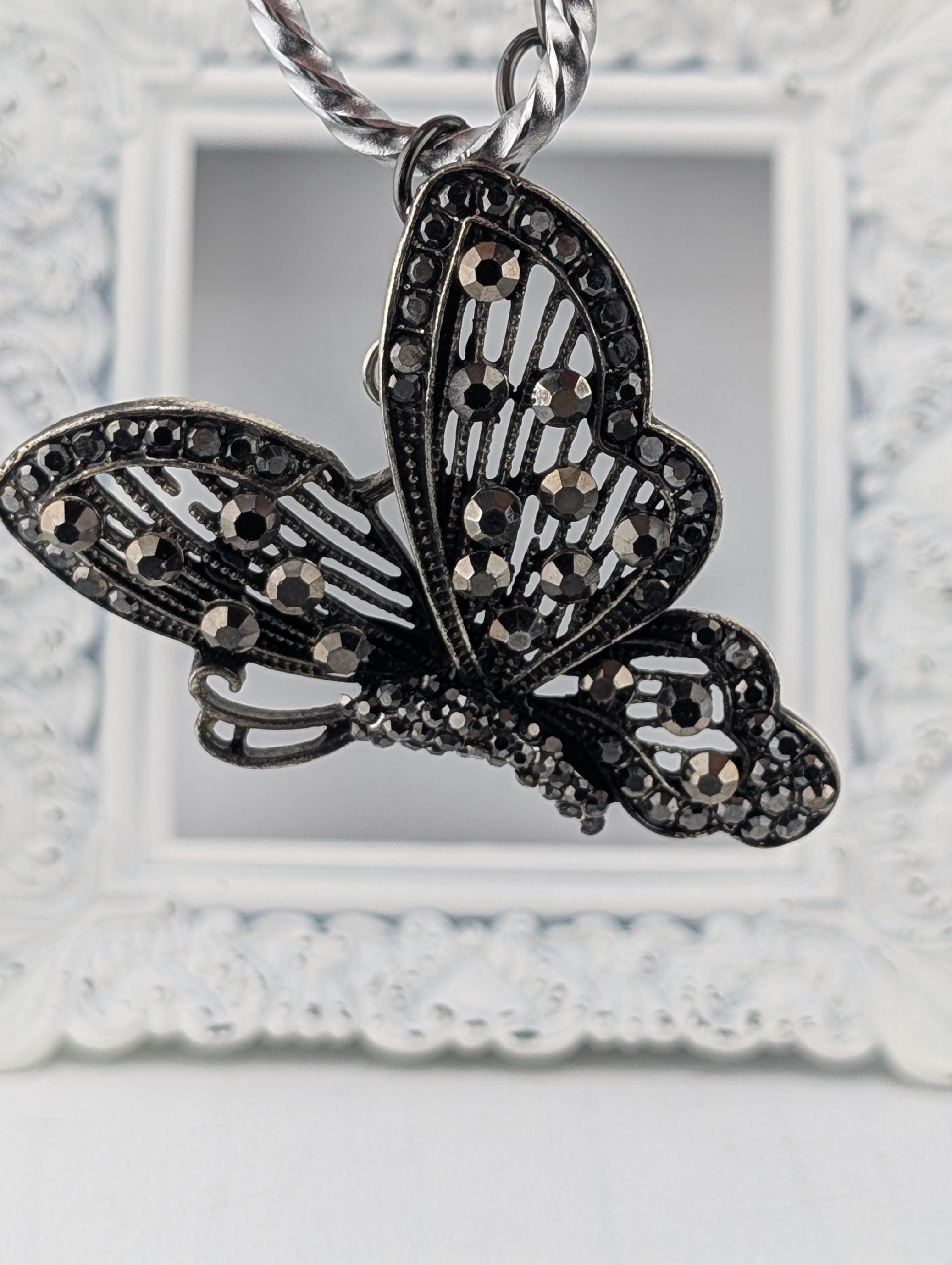 A decorative butterfly pendant with intricate black and silver designs, including rhinestones or crystals, hanging from a chain.