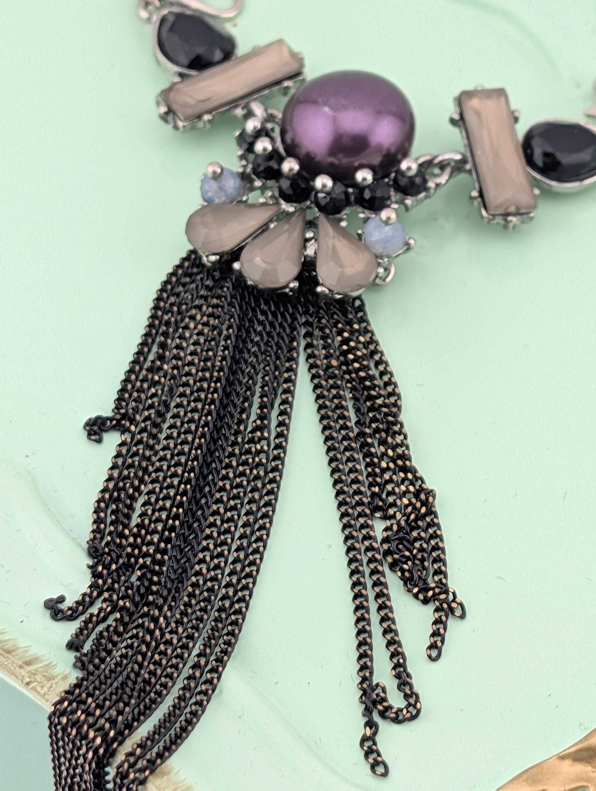 A close-up image of a necklace with a purple gemstone and a long, black tassel.