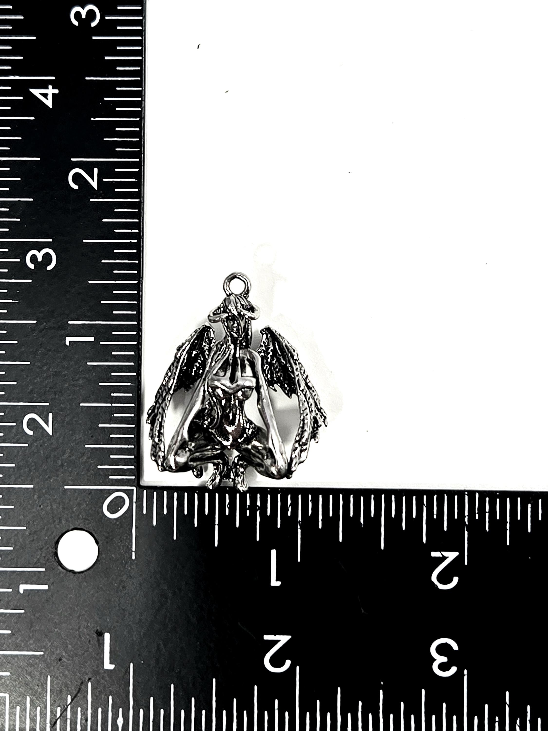 A silver-colored pendant or charm is placed next to a ruler, indicating its size.