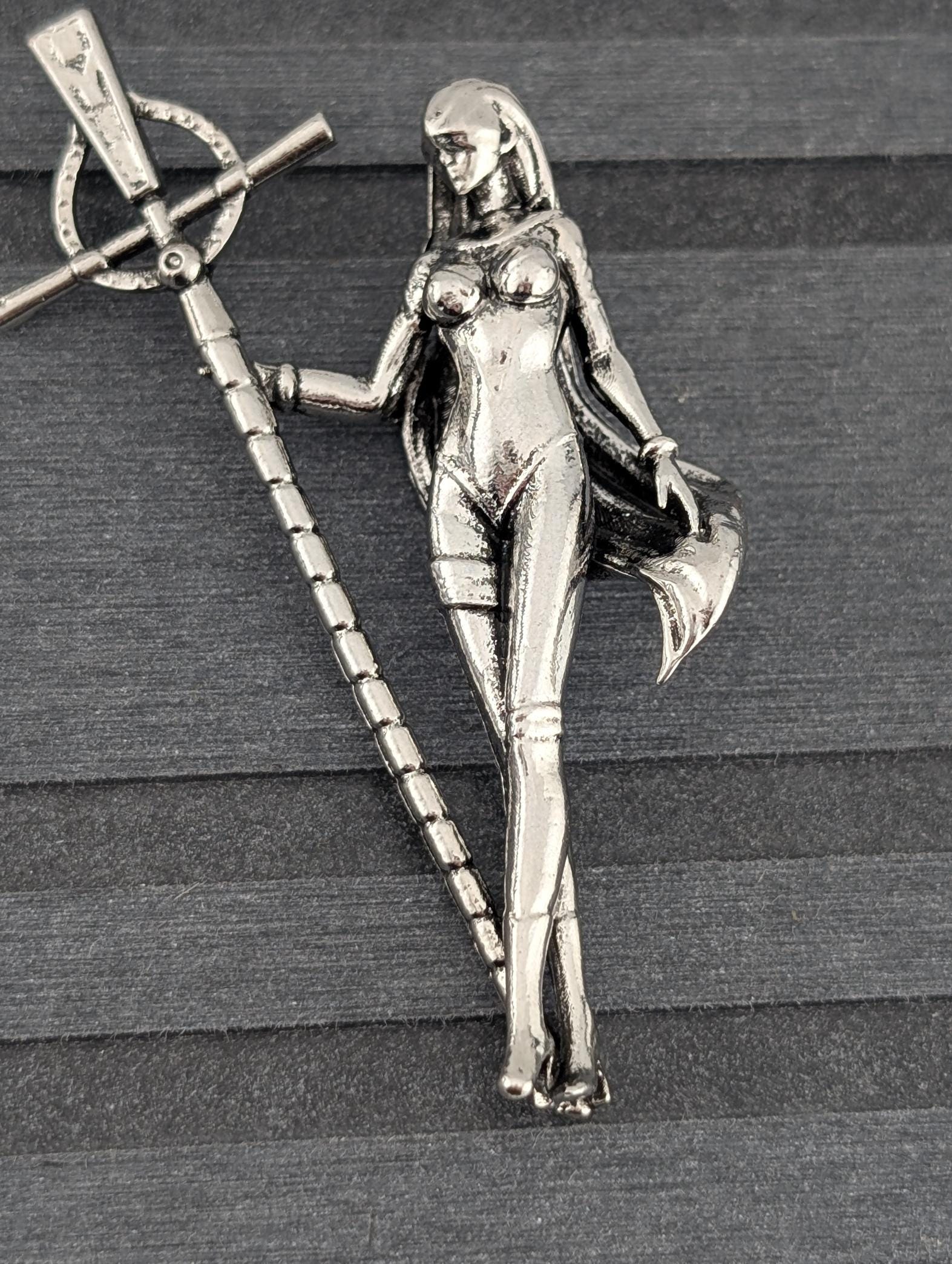 A silver-colored metal brooch in the shape of a female figure, holding a staff or staff-like object.