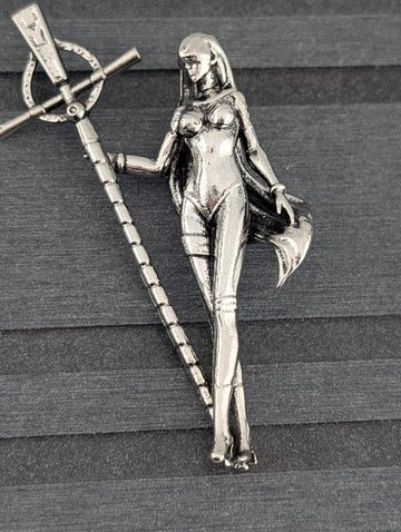 A silver-colored metal brooch in the shape of a female figure, holding a staff or staff-like object.