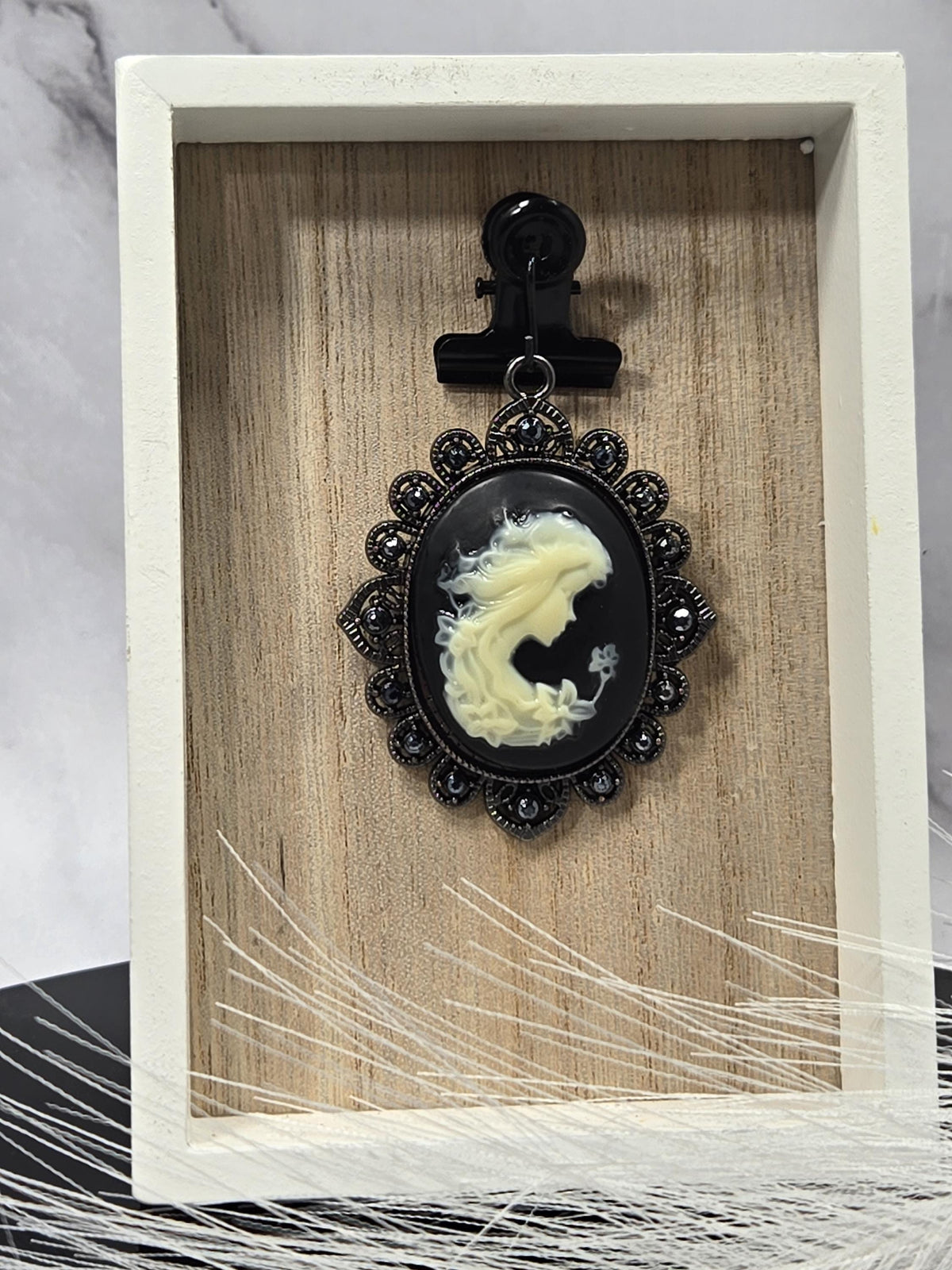 a black oval brooch with a white design, placed on a wooden surface inside a white frame.