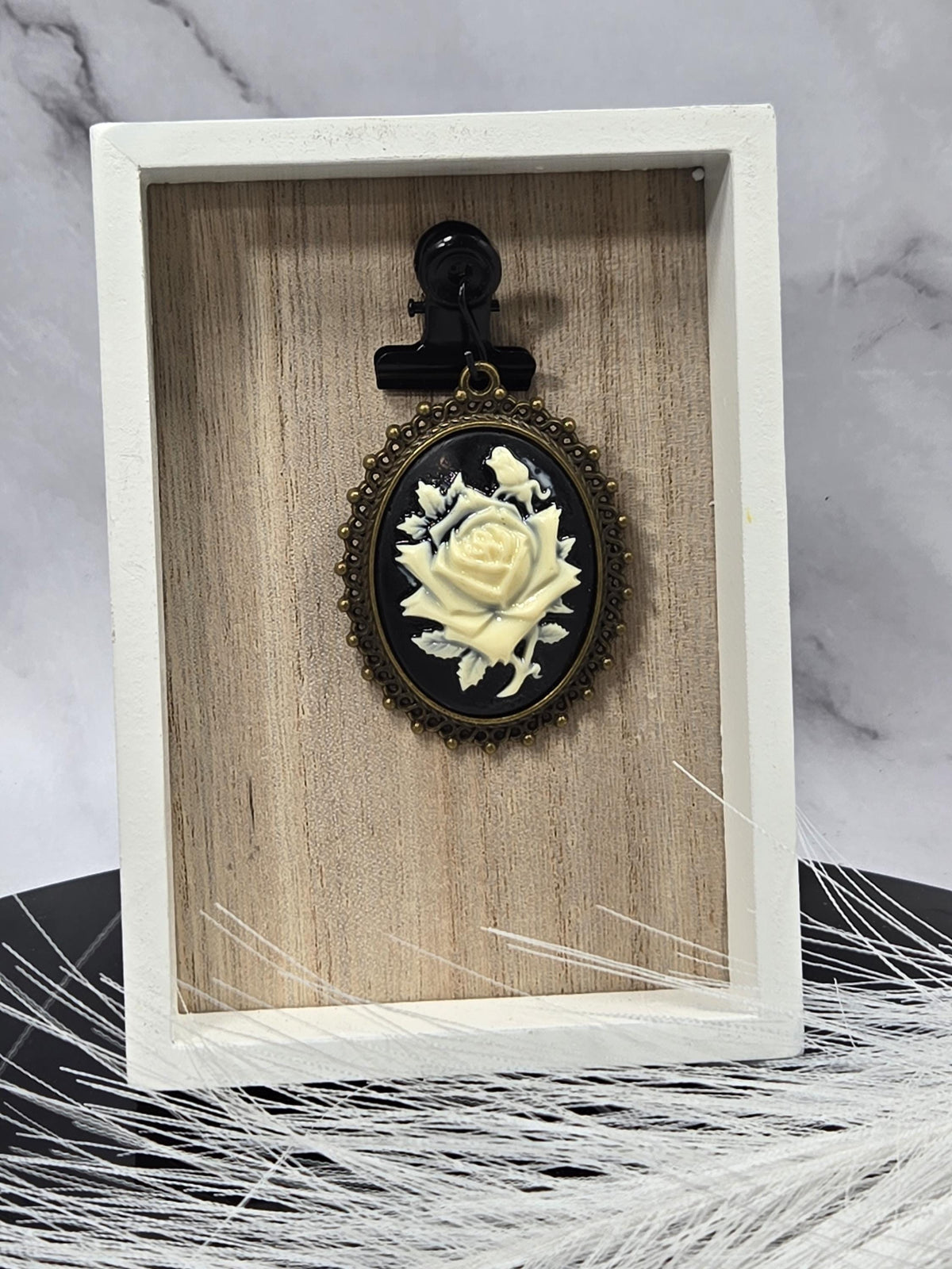 A black oval pendant with a white rose design is displayed in a white wooden frame.
