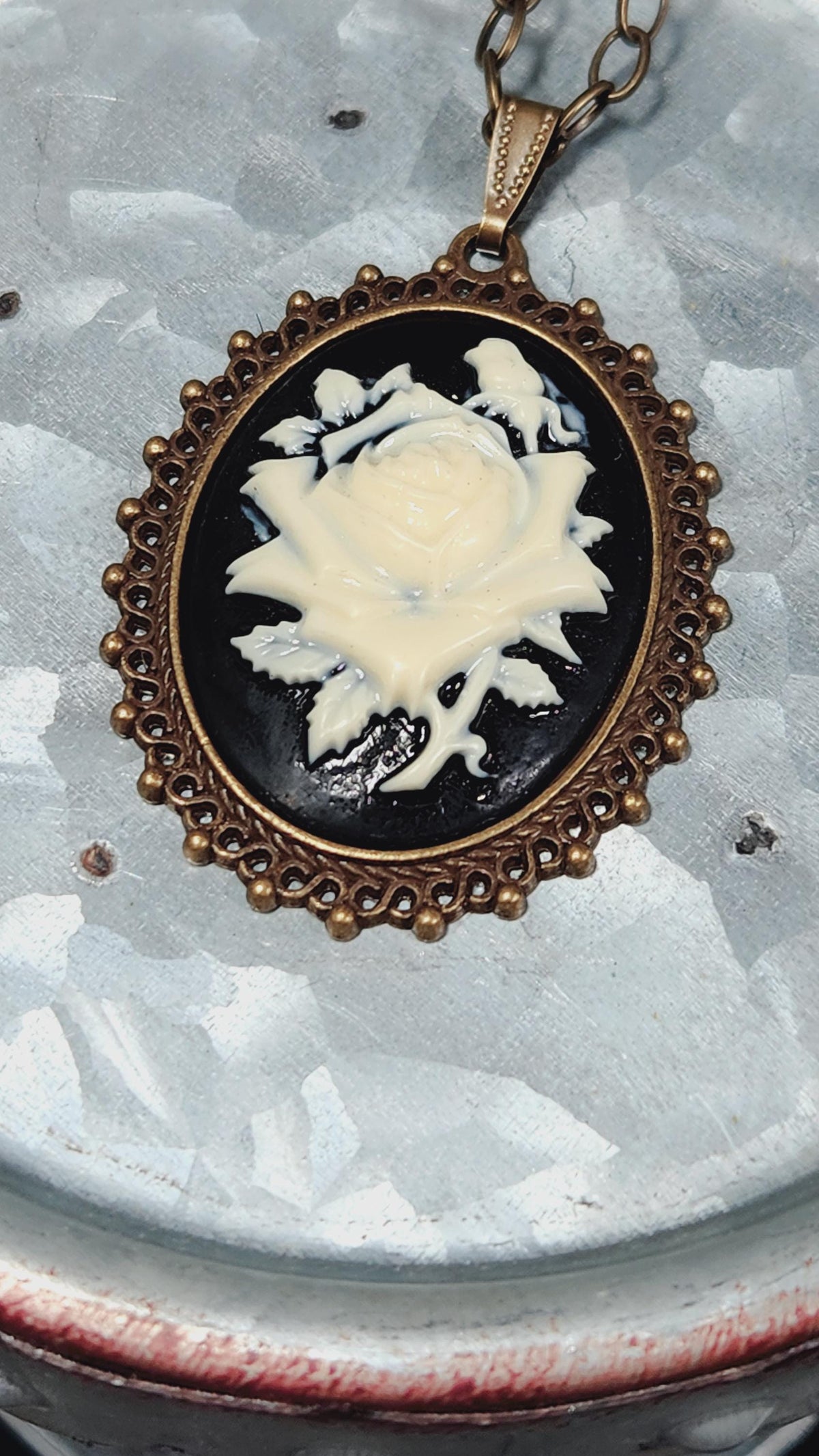 A decorative pendant featuring a white rose design on a black background, encircled by an ornate gold frame.