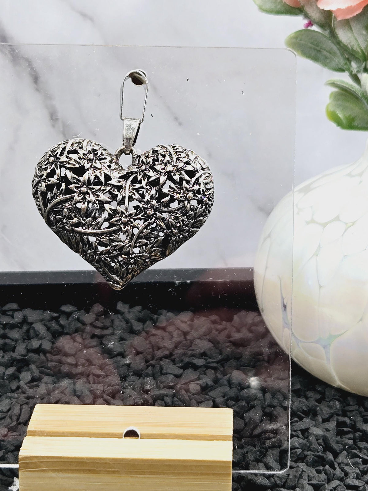 a heart-shaped metal object with a floral design, placed on a marble surface next to a white vase and a wooden object.
