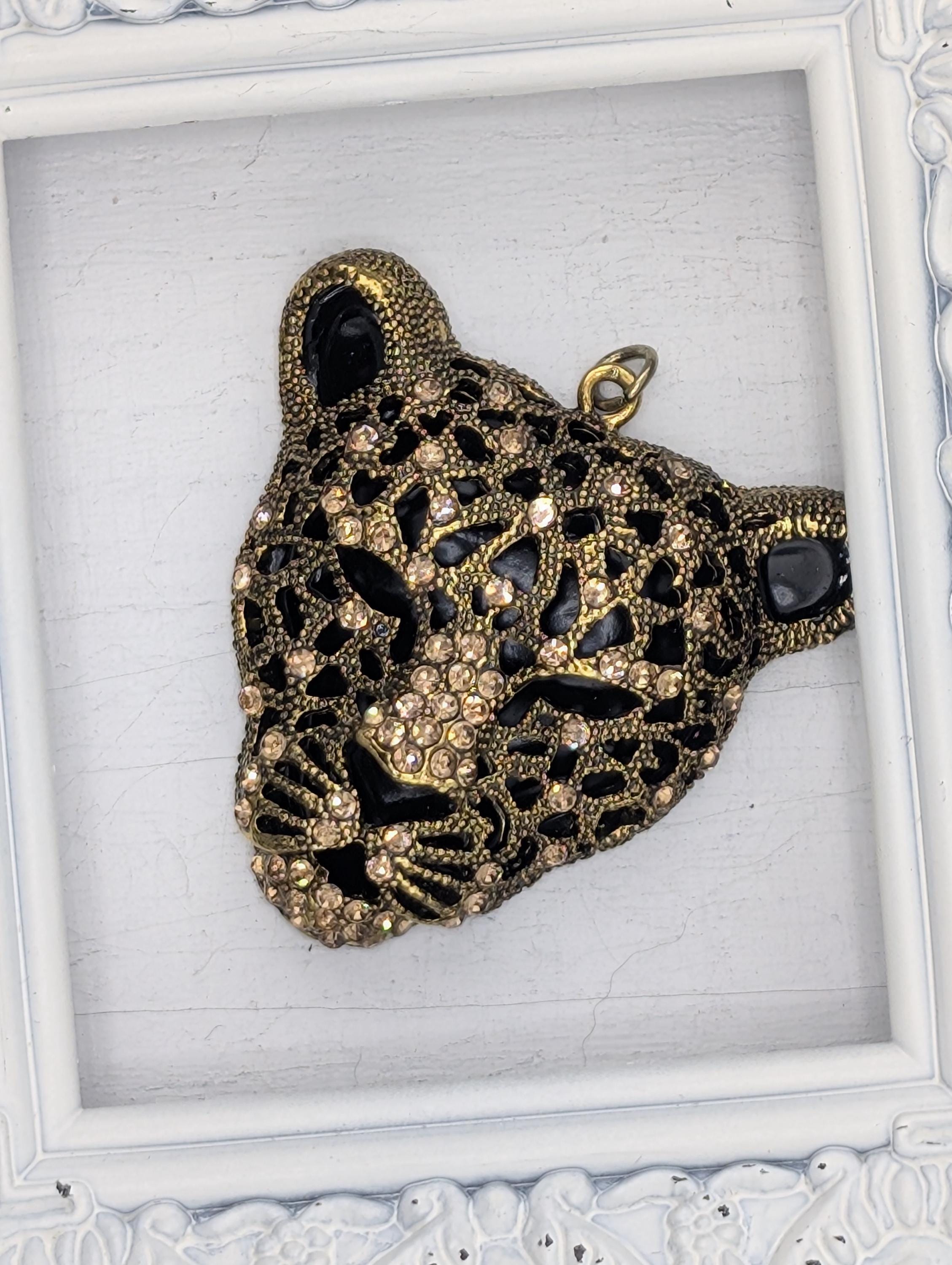 A decorative pendant in the shape of a leopard's head, adorned with intricate gold and black designs and sparkling rhinestones.
