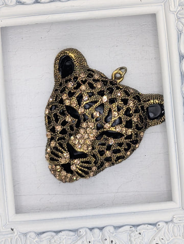 A decorative pendant in the shape of a leopard's head, adorned with intricate gold and black designs and sparkling rhinestones.