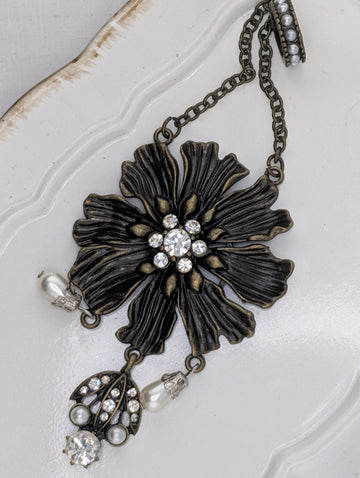 A decorative piece of jewelry featuring a large, intricately designed flower-shaped pendant with a central stone surrounded by smaller stones. The pendant is attached to a chain with pearls hanging from it, creating a stylish and elegant accessory.