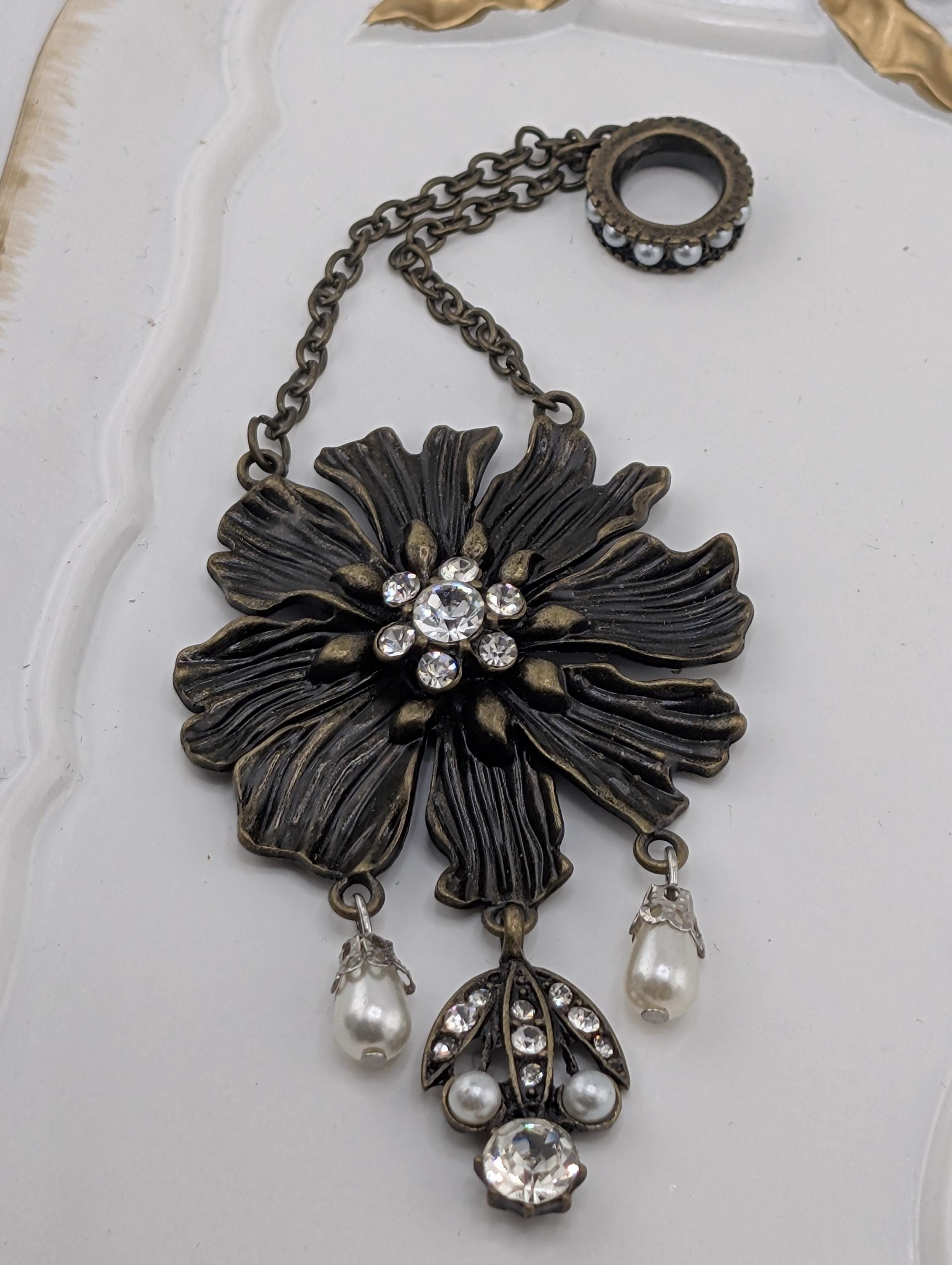 A decorative pendant featuring a large, intricately designed flower with a central stone surrounded by smaller stones, pearls, and dangling charms.