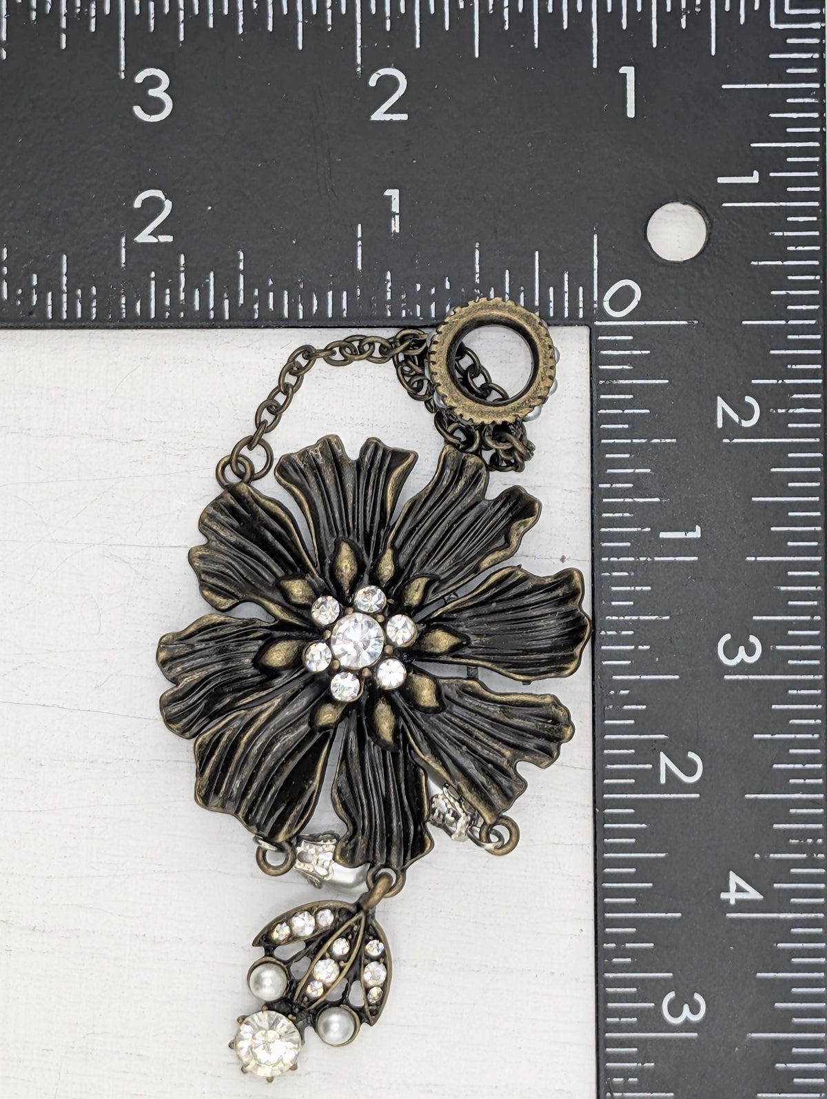 A close-up image of a decorative piece with a large, intricate flower-like design and sparkling rhinestones or crystals. The piece is placed next to a ruler for size comparison.