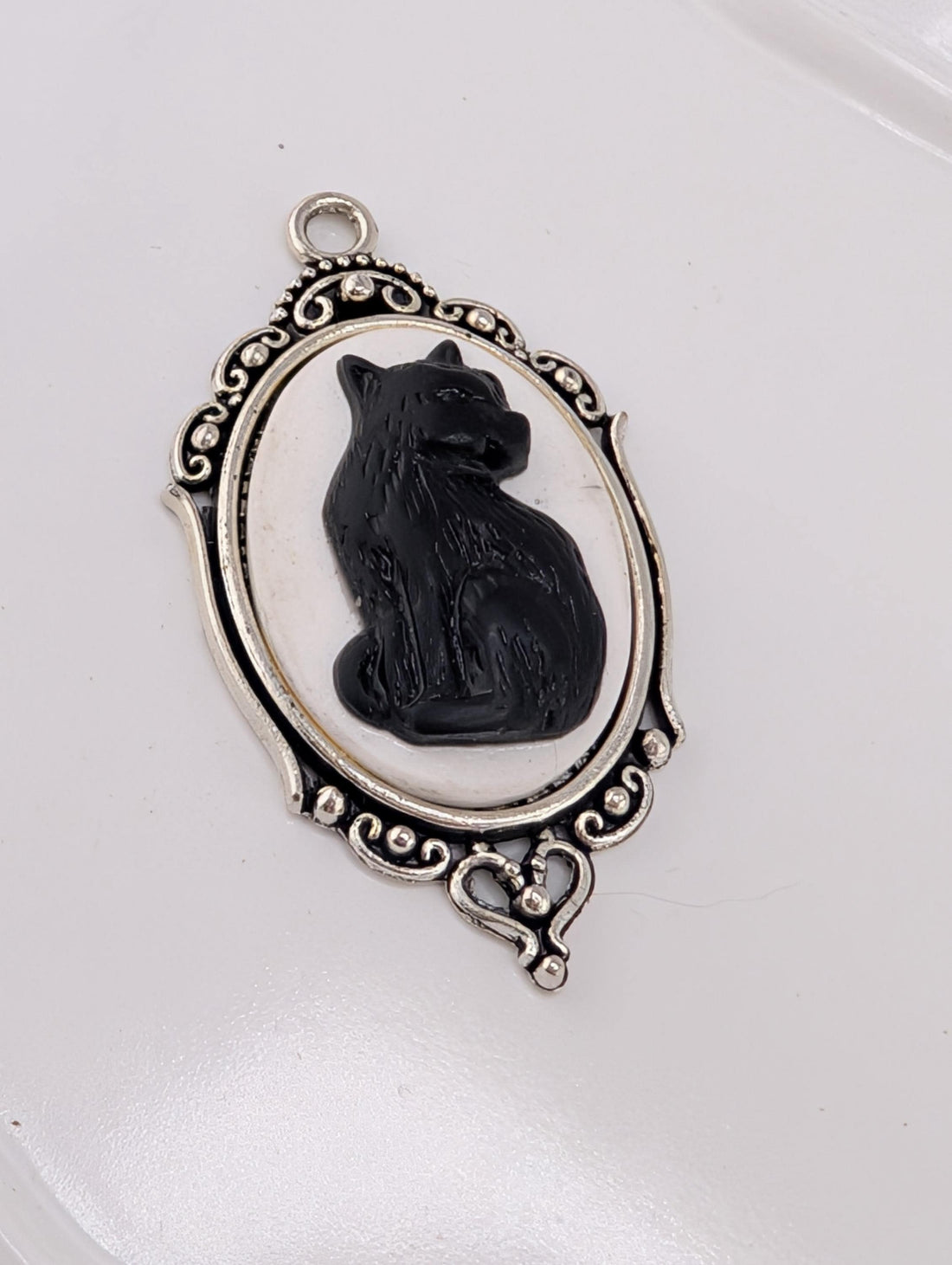 A silver-toned pendant featuring a black cat sitting inside an oval frame with an ornate border.