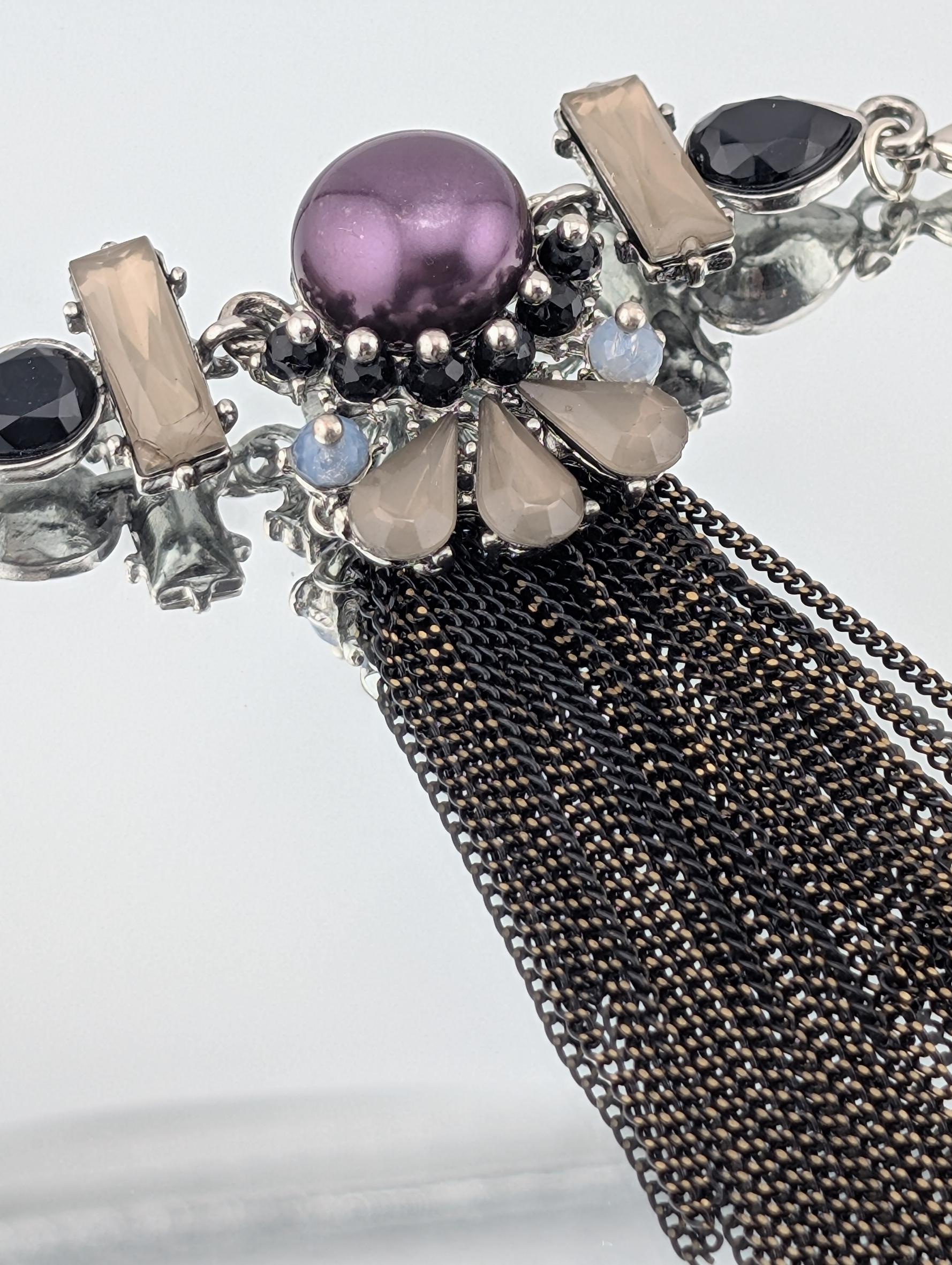 A close-up view of a decorative necklace with a combination of purple, black, and white gemstones, as well as a tassel made of black and gold chain.