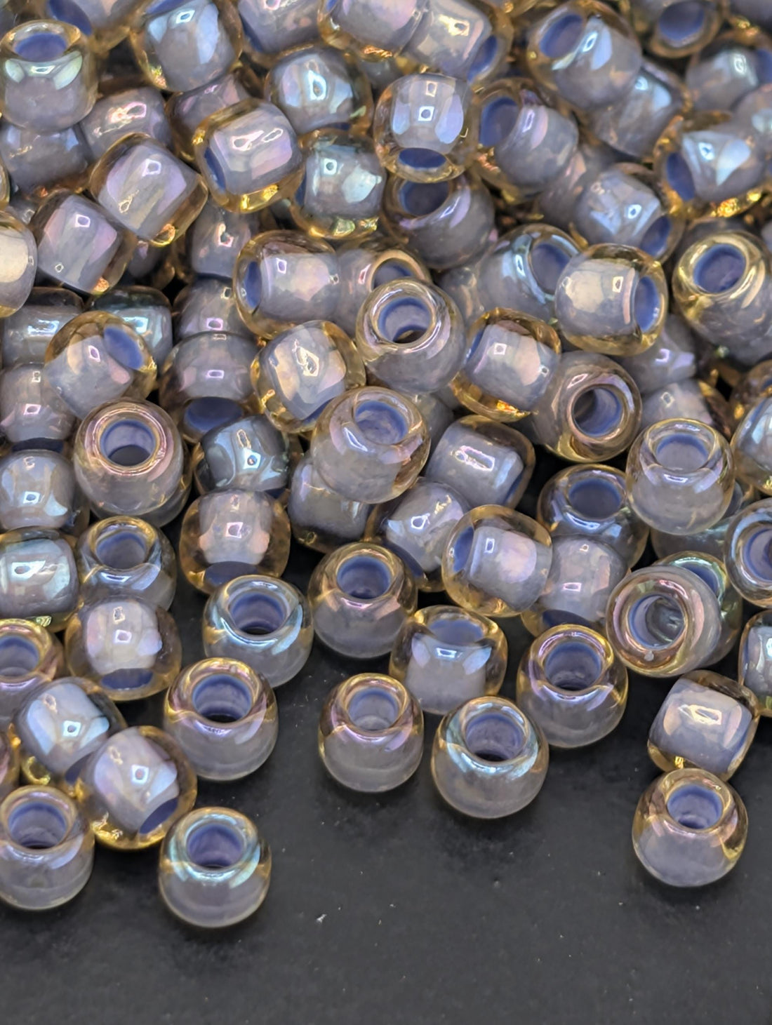 a close-up view of numerous small, clear beads scattered across a dark surface. The beads appear to be made of a translucent material, possibly glass or plastic, and they come in various colors, including shades of blue, yellow, and purple. The arrangement of the beads creates a visually interesting and textured composition.