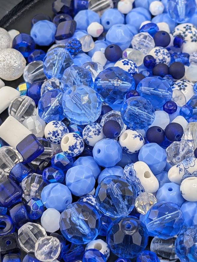 The image is a close-up of various blue and white beads, including clear and white beads, as well as blue and white beads with floral designs. The beads are scattered and overlapping, creating a visually interesting and colorful composition.