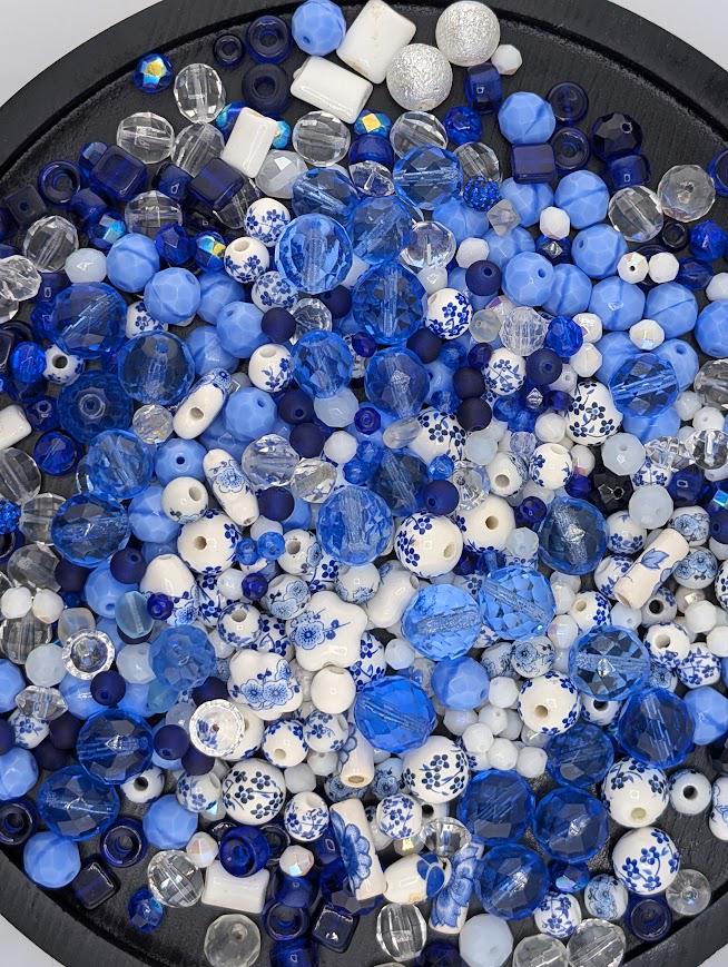 The image is a collection of various colored glass beads, including blue, white, and clear beads, arranged on a black surface.