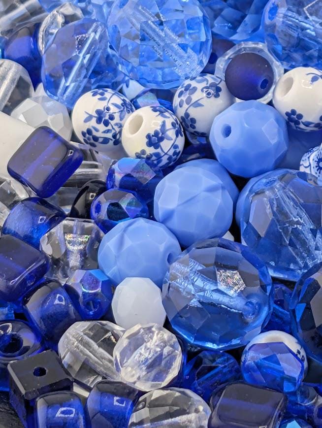The image is a close-up of various blue and white glass beads, including clear and blue glass beads, as well as some white glass beads with blue floral designs. The beads are scattered and overlapping, creating a visually interesting and colorful composition.