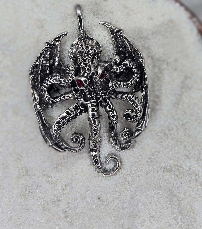 A silver pendant in the shape of a winged creature with intricate details and a red gemstone.
