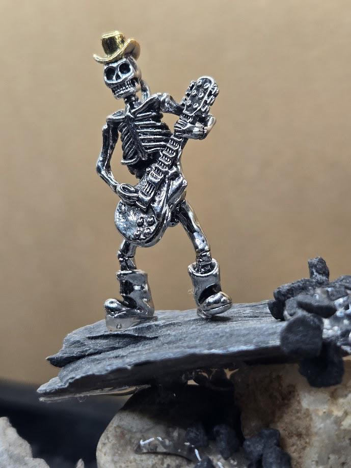 A metal skeleton wearing a hat and playing a guitar is standing on a rock formation.