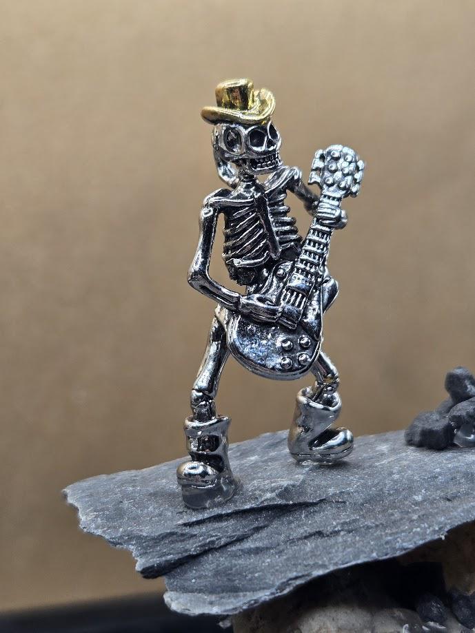 A small metal figurine of a skeleton playing a guitar, wearing a hat, and standing on a rock.