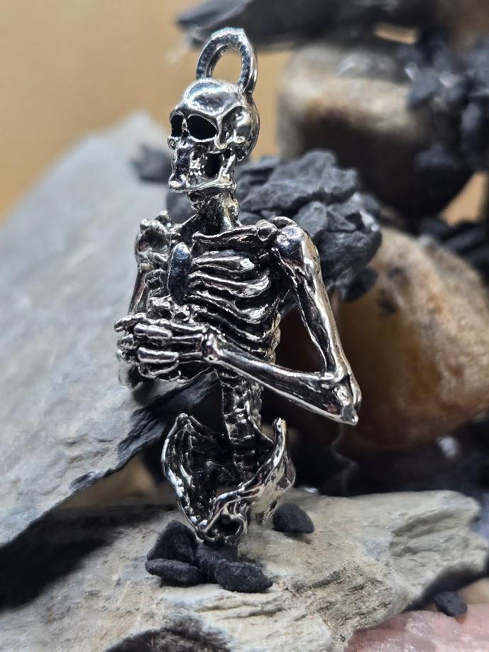 A silver-colored metal skeleton pendant is displayed against a backdrop of rocks and other natural elements.
