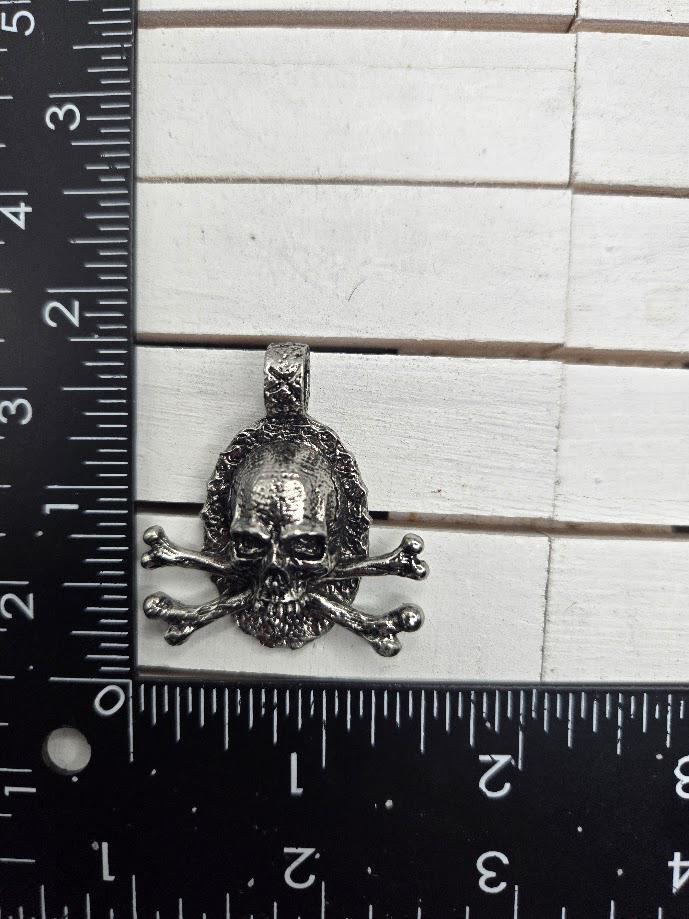 A silver-colored pendant in the shape of a skull with crossed bones, placed next to a ruler for size comparison.