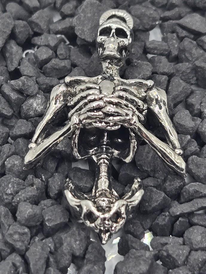 A silver-colored metal skeleton pendant is shown against a background of small rocks.