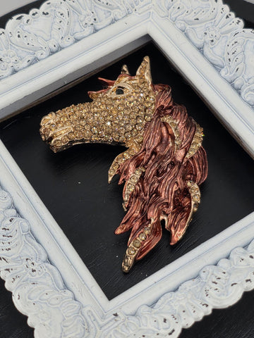 A decorative piece featuring a golden horse head with intricate details and sparkling rhinestones, displayed within a white ornate frame.