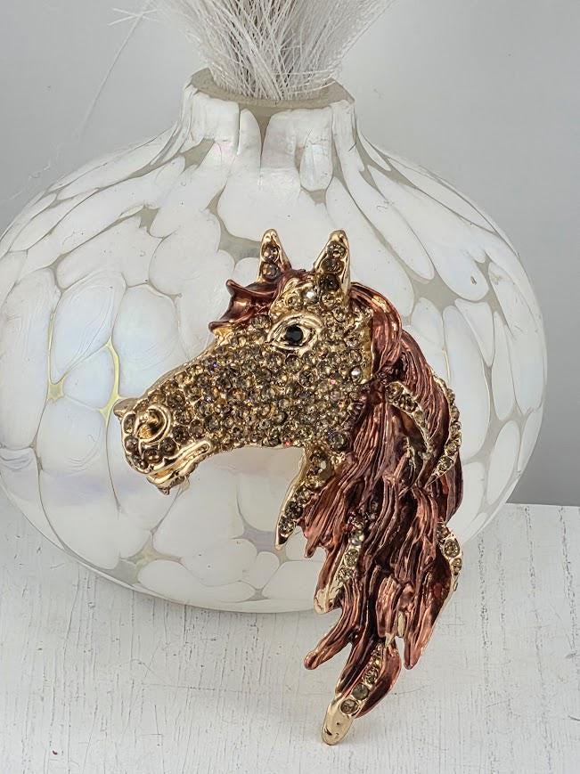 A decorative horse head brooch with a gold and brown color scheme, placed on a white vase with a white floral pattern.