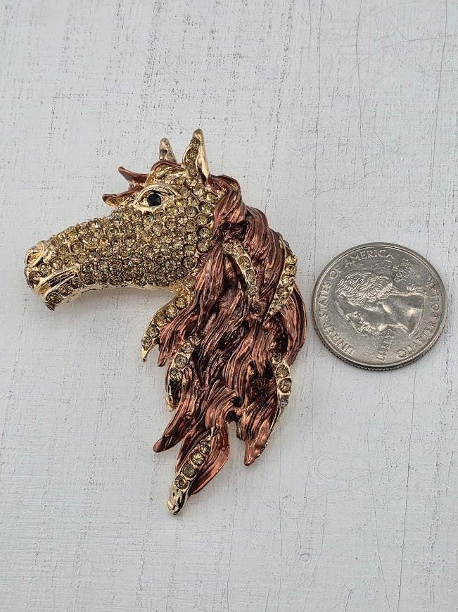 A decorative horse head brooch made of gold and adorned with rhinestones is placed next to a United States quarter.