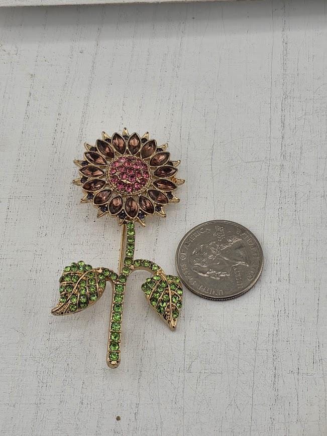 A decorative brooch in the shape of a sunflower, adorned with green gemstones, is placed next to a United States quarter coin on a white surface.