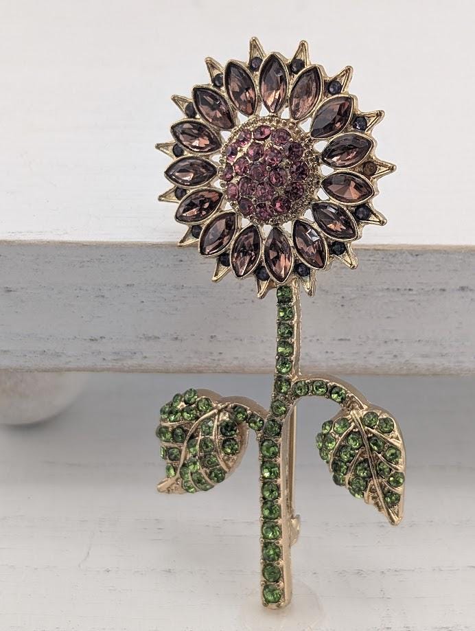 A decorative brooch in the shape of a sunflower, adorned with green gemstones and purple gemstones.