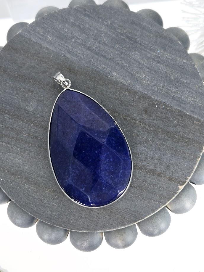 A large, blue gemstone pendant with a silver frame is displayed on a white surface.