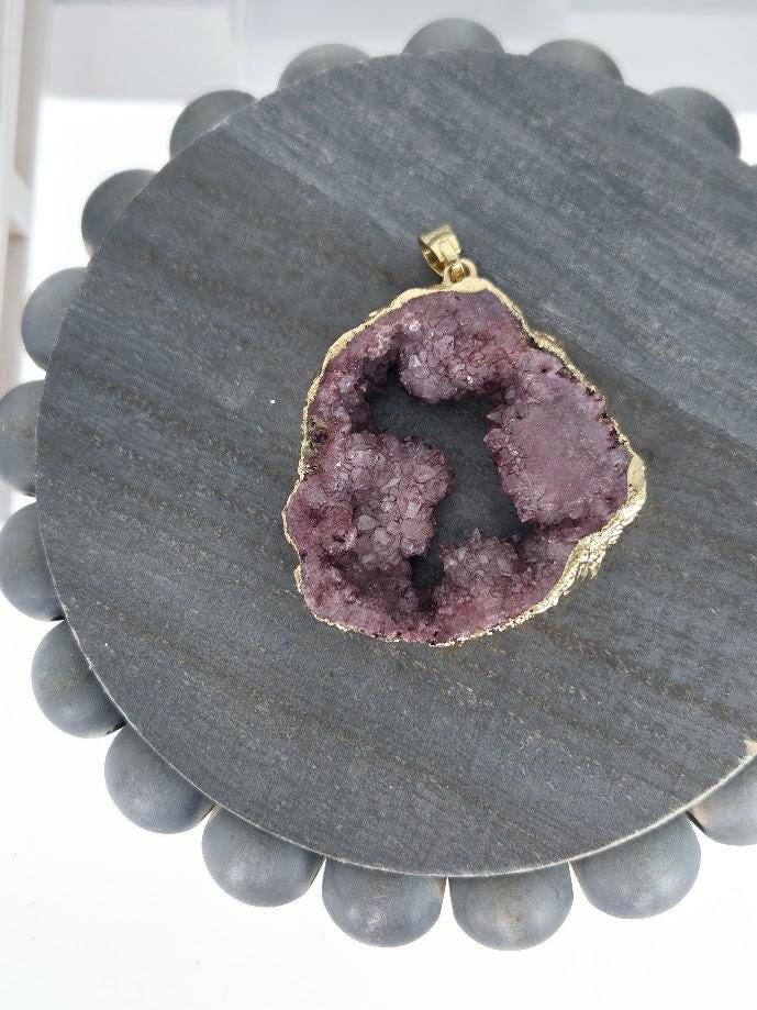 A large, purple geode pendant with a gold-colored frame is displayed on a gray circular stand.